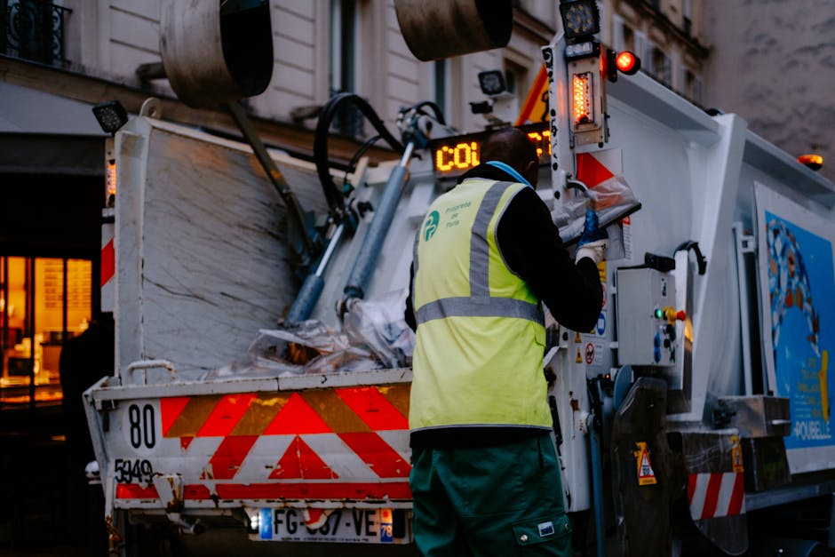 A waste collection vehicle equipped with hydraulic lifting mechanisms is parked outdoors during dusk, with warm interior lighting visible through nearby windows. The vehicle's rear compartment is open