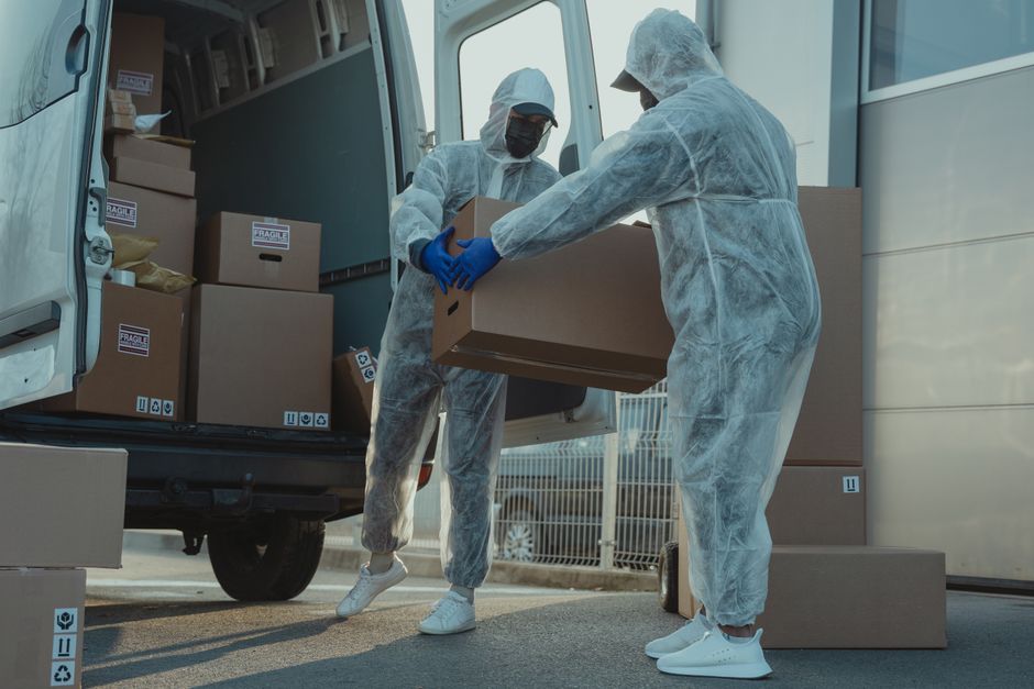Two movers dressed in white protective suits, face masks, and blue gloves are loading cardboard boxes into the back of a white moving van parked near a building on Electric Avenue. The open van reveal